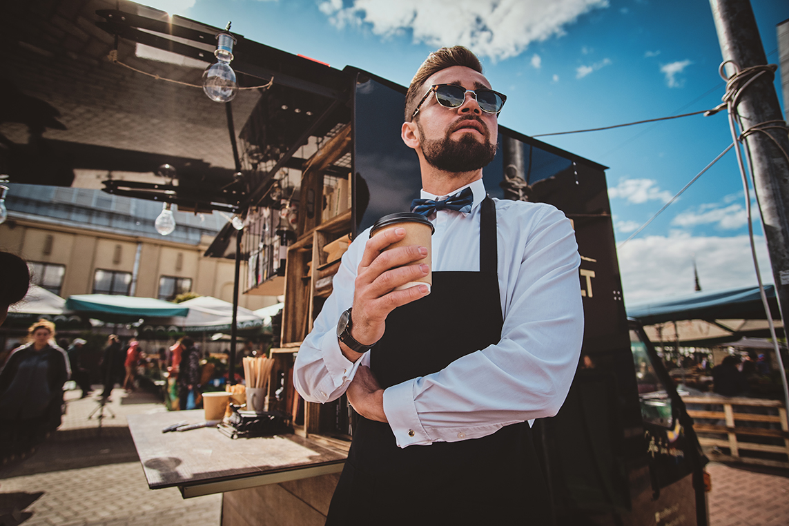 Groomed trendy barista in glasses has a coffee break and enjoying coffee. There are bright sunny summer day.